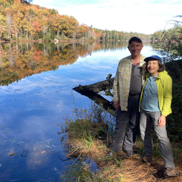 Tim and Di standing at the edge of a perfectly reflective lake in the fall, smiling at the camera.