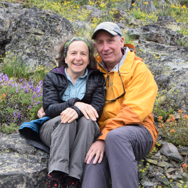 Tim and Di sitting together on a rock surrounded by mountain wildflowers, both smiling warmly at the camera.